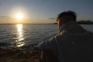 A man sitting by a calm lake at sunset, reflecting on techniques for how to be less emotionally reactive.
