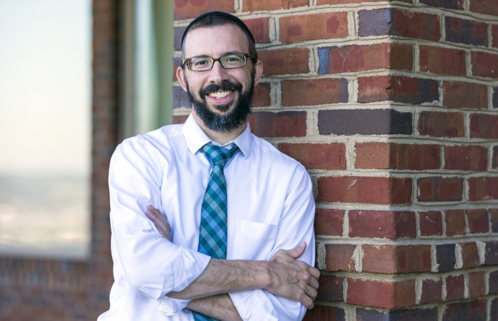 Mindfully clinician smiling confidently while leaning against a brick wall, wearing a white shirt and blue plaid tie.