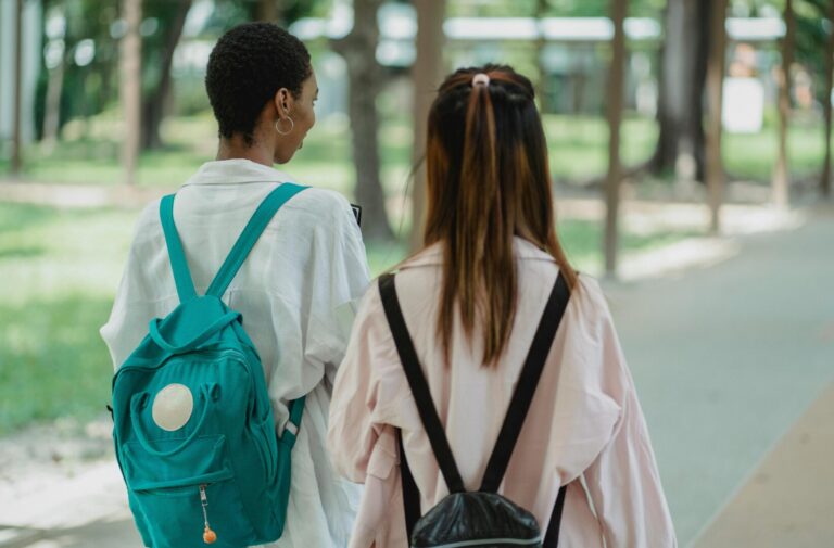 Two diverse female students with backpacks walking together on a school campus, representing social support and peer connection.