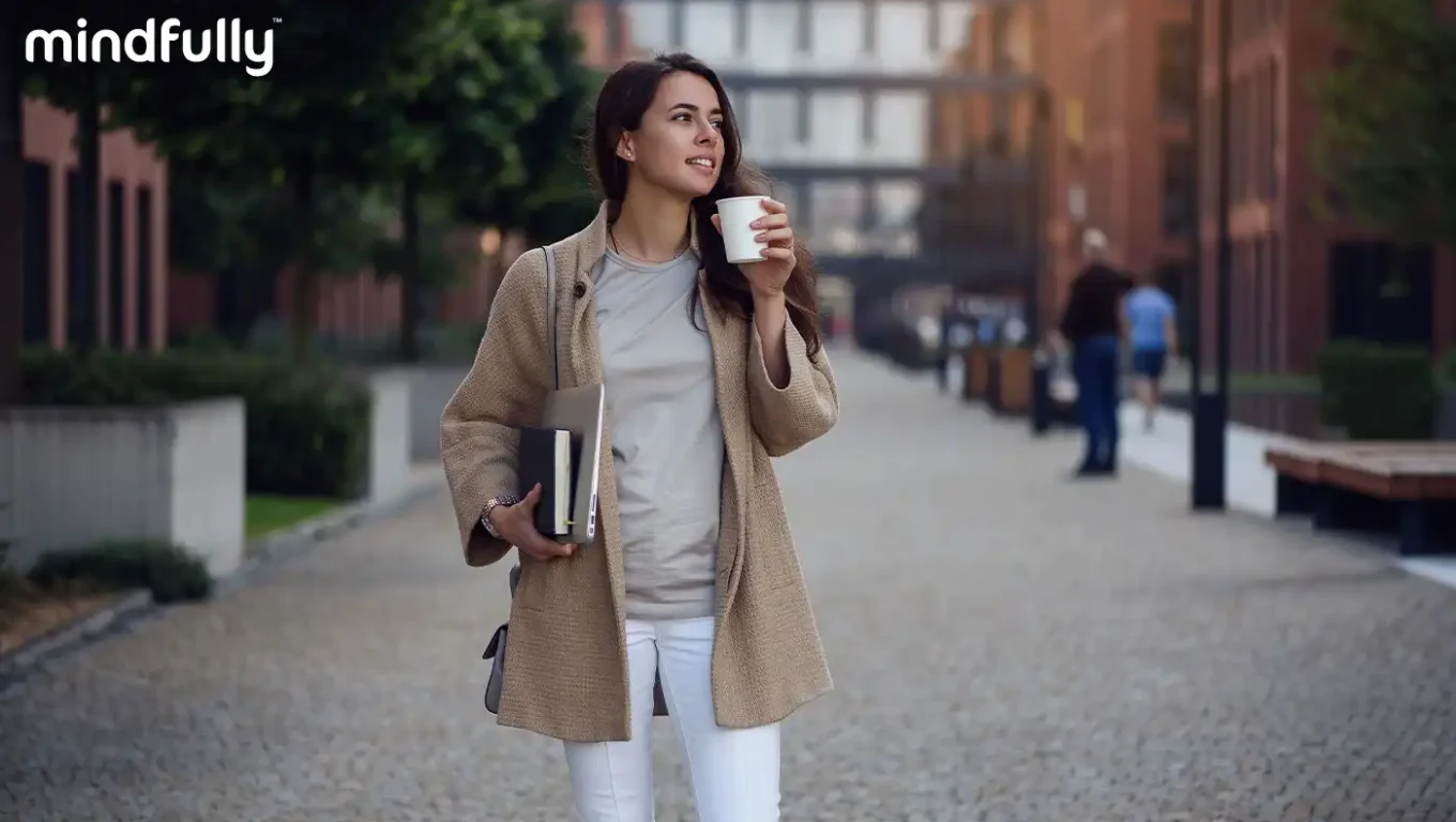 Woman holding coffee and notebook while walking calmly through urban environment, representing mindful stress management and self-care during work challenges