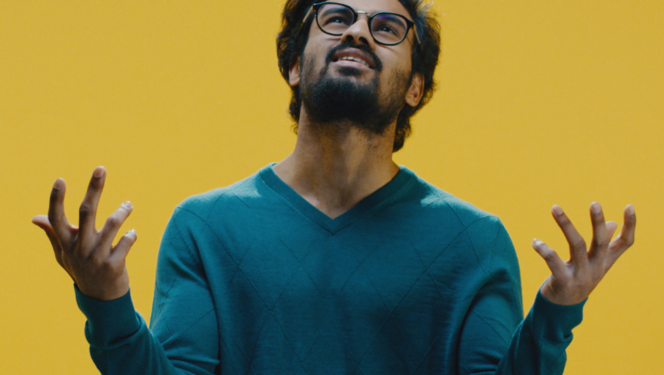 A man in a green sweater and glasses stands before a cheerful yellow background in a anger mode.