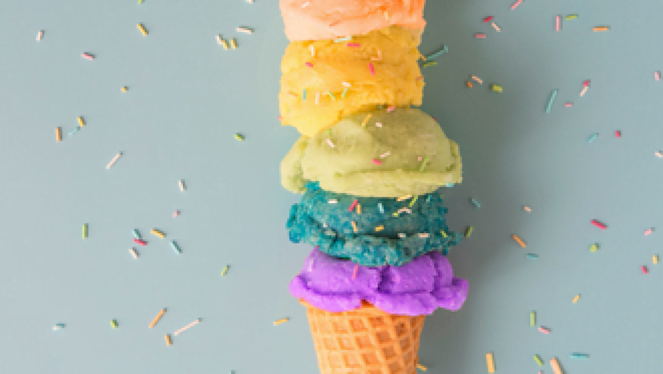 A vibrant rainbow ice cream cone topped with colorful sprinkles against a neutral background.